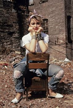 a woman sitting on top of a wooden chair in front of a brick building with her hands to her face