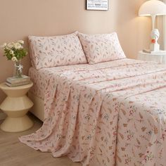 a bed covered in pink floral sheets and pillow cases next to a lamp on a table