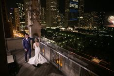 a bride and groom standing on a balcony overlooking the city at night with fireworks in the sky