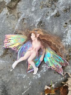 a fairy doll is laying on the ground next to leaves and rocks with her hair blowing in the wind