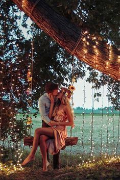 a man and woman sitting on a bench under a tree with fairy lights around them