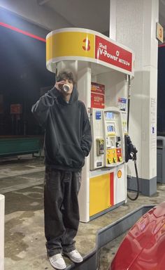 a man standing in front of a gas pump drinking from a cup while holding a cell phone up to his ear