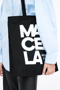 A canvas bucket tote bag in 100% European cotton, with a bottom panel for added capacity. The tote features an oversized MARCELLA NEW YORK graphic on one side, in white print on black canvas.[SPLIT] Our production team enjoys a living wage, a 40-hour work week with paid overtime, free health insurance and 20 days annual paid vacation. Black Rectangular Canvas Bag For Weekend, Casual Black Bags With Graphic Design, Large Capacity Black Cotton Canvas Bag, Cotton Tote Shoulder Bag With Logo Print, Black Canvas Weekend Bag, Large Capacity Cotton Bucket Canvas Bag, Black Canvas Bag For Weekend, Black Canvas Bag For The Weekend, Black Rectangular Canvas Bag With Graphic Print