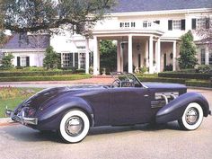 an old purple car parked in front of a large white house with trees and bushes