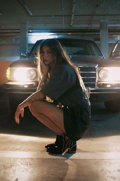 a woman squatting on the ground in front of a car with her legs crossed