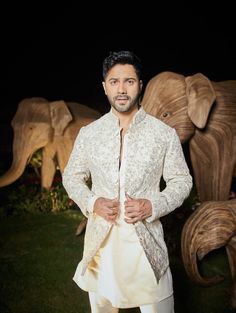 a man standing in front of an elephant statue wearing a white suit and jacket with his hands on his hips