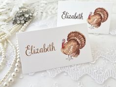 two place cards with turkeys on them sitting on a lace tablecloth next to pearls