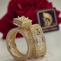 two gold wedding rings sitting next to each other on top of a white surface with a red rose in the background