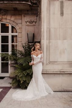 a woman in a white wedding dress standing next to a building with greenery on it