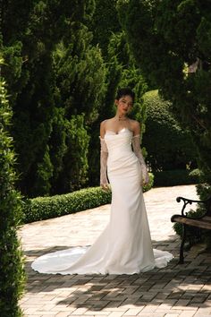 a woman in a white wedding dress standing on a brick walkway next to a park bench