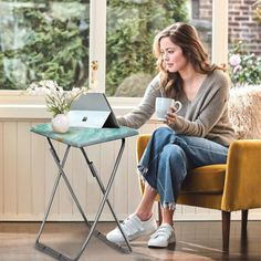 a woman sitting in a chair holding a coffee cup and looking at her laptop computer