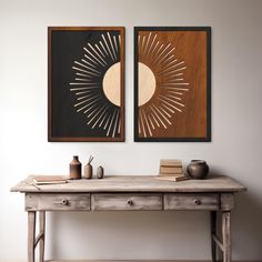 two framed art pieces on the wall above a wooden table with books and vases