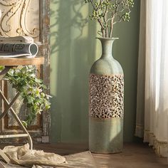 a green vase with flowers in it sitting on a table next to a chair and window