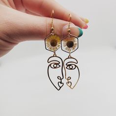 a woman's hand holding two earrings with sunflowers on them and a face in the middle