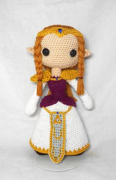 a crocheted doll wearing a purple dress and yellow headband, standing in front of a white backdrop