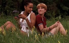 two women sitting in the grass looking at their cell phones and smiling for the camera