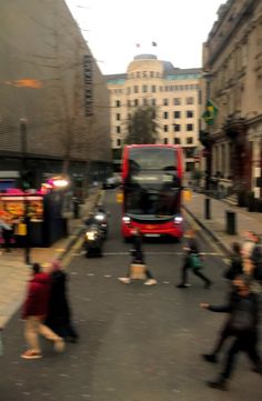 a red double decker bus driving down a street next to people walking on the sidewalk