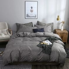 a bed with grey linens and pillows in a white room next to a chair