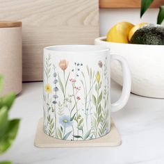 a white coffee mug sitting on top of a counter next to a bowl of fruit