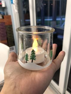 a hand holding a small glass candle holder with trees in it and snow on the ground