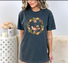 Introducing our charming country style farmhouse fall wreath with mushrooms and moth tshirt! This unique and whimsical tee captures the essence of cozy autumn vibes in an instant. It features an enchanting arrangement of earthy mushrooms and an elegantly detailed moth at the center, adding a touch of nature's beauty to your fall wardrobe. With its rustic farmhouse style and warm color palette, this shirt is the perfect addition to your fall  collection. Enjoy this warm addition to your everyday comfort wear while embracing the spirit of the season with our delightful country style farmhouse fall wreath tshirt. Comfort Colors introduces its garment-dyed t-shirt; a fully customizable tee made 100% with ring-spun cotton. The soft-washed, garment-dyed fabric brings extra coziness to your wardr Cottagecore Short Sleeve T-shirt For Fall, Fall Cottagecore Tops With Graphic Print, Fall Mushroom Design Crew Neck T-shirt, Cottagecore Graphic Print Tops For Fall, Fall Crew Neck T-shirt With Mushroom Design, Cotton T-shirt With Mushroom Design For Fall, Fall Mushroom Print Graphic Tee, Fall Mushroom Print Graphic T-shirt, Fall Graphic Tee With Mushroom Print