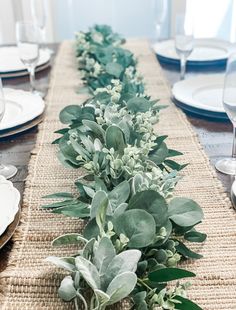 the table is set with white plates and place settings, along with greenery on each side