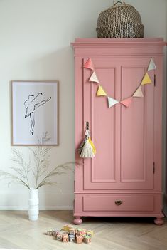 a pink armoire next to a white vase with flowers in it and some toys on the floor