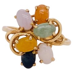 A depth of color and layers are the beauty of this ring. All of the jades in this ring are naturally occurring colors (white, black, yellow, lavender, orange, green). The everyday strength of jade often gives a source of strength for any who need it. A wire work frame is securely layered to hold each of the colorful jade cabochons along with swirling scroll work accents near the shoulders of the band. The ring top design forms a circle that measures 17.5 millimeters in diameter. This charmer was made in Hong Kong (stamped: 585 Hong Kong 14K) and is built to be slender yet strong. The full details for this beautiful ring are listed below: Metal Quality: 14 karat yellow gold Complete Ring Weight: 3.7 grams Ring Size: 8.25 (sizing not included) Band Width: Center design 17.5 mm in diameter, s Fine Jewelry Multicolor Cabochon Gemstones, Multicolor Cabochon Gemstones For Fine Jewelry, Multicolor Cabochon Gemstones Fine Jewelry, Multicolor Cabochon Rings In Fine Jewelry Style, Multicolor Cabochon Rings Fine Jewelry, Multicolor Oval Rings With Natural Stones, Vintage Cluster Ring, Lavender Orange, Scroll Work