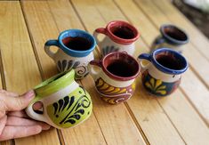 a hand holding a coffee mug next to several different colored cups on a wooden table