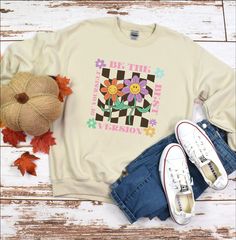 "Who doesn't love a fun retro inspired sweatshirt? Our \"Be the Best Version of Yourself\" sweatshirt is so comfortable, you'll want to wear it all the time! Full of fun vintage vibes, this is a wardrobe essential. With a checkerboard background and happy daisies design, this shirt will quickly become everyone's fave. Available in many sizes and colors. The shirt is made from medium-heavy weight cotton/polyester blend and is soft and comfortable, making it perfect for all day wear. With its unique design and inspirational message, you will make a statement every time you pull this sweatshirt on. **Be sure to review your order before completing your purchase. All orders are sent directly into production, therefore cancellations are not possible, and refunds are not accepted.**" Checkerboard Background, Happy Daisy, Retro Sweatshirts, Best Version Of Yourself, Sweatshirt Vintage, Trendy Shirts, Inspirational Message, Be The Best, Vintage Vibes