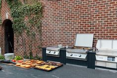 an outdoor bbq grill with food on the table next to it and a brick wall behind it