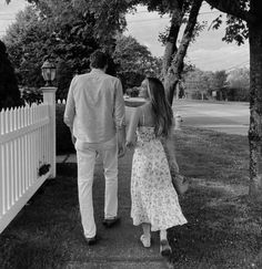 a man and woman walking hand in hand down a sidewalk next to a white picket fence