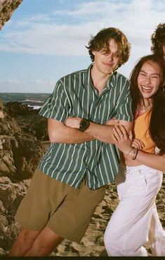 two young people standing next to each other on a rocky beach with the ocean in the background