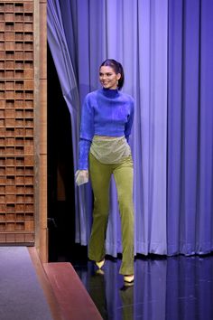 a woman standing in front of a purple curtain wearing green pants and a blue sweater