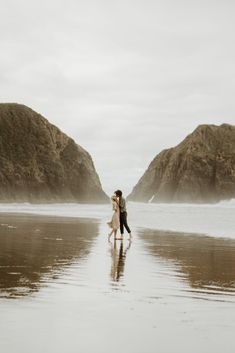 a man and woman standing on top of a beach next to the ocean with mountains in the background