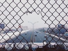 the view from behind a chain link fence looking down at a street with cars driving on it