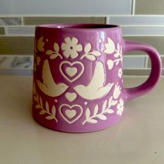 a purple coffee mug with white birds and hearts painted on the side, sitting on a counter