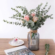 a glass vase with flowers and greenery on a table next to an open magazine