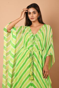 Lime green kaftan tunic with all-over Leheriya print, embroidered neckline and criss-cross tie-up. Comes with braided belt.
Component: 2
Printed, Embroidered
Neckline: V-Neck
Sleeve Length: Asymmetric
Fabric: Georgette
Color: Green
Asymmetric hem
Braided belt with tassels
Note: The inner slip worn by the model is not for sale - Aza Fashions Traditional Green V-neck Tunic, Green V-neck Kaftan For Vacation, Green V-neck Free Size Kaftan, Summer Festive V-neck Kaftan, Navratri Dress With Back Tassel Tie-up, Green Tunic Kaftan For Festive Occasions, Green Bohemian Tunic Kaftan, Green V-neck Kaftan For Beachwear, Festive Green V-neck Kaftan