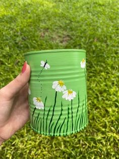 a hand holding a green tin can with daisies painted on it in the grass