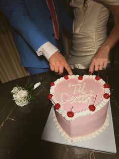 a man and woman cutting into a pink heart shaped cake with cherries on it