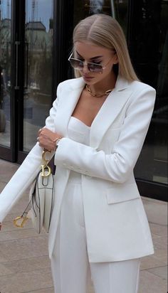 a woman in white suit and sunglasses walking down the street with her hand on her purse