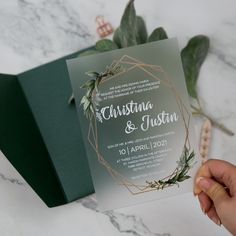 a person holding up a wedding card in front of a box with greenery on it