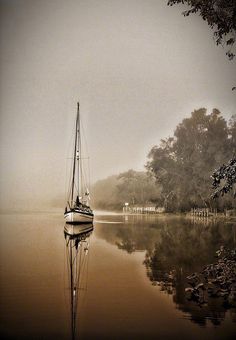 a sailboat floating on top of a lake next to a shore covered in fog