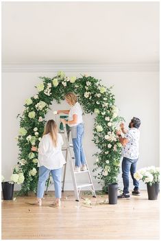 two women are decorating a floral wreath with white flowers and greenery on the wall