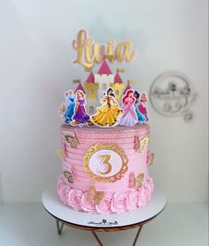 a three tiered cake with princess figures on top and pink icing, sitting on a table