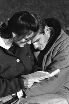 a man and woman sitting next to each other on the ground looking at a book