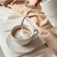 a person pouring milk into a cup on top of a white saucer next to a book