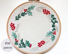 an embroidered christmas wreath with holly leaves and red berries on white fabric in a hoop