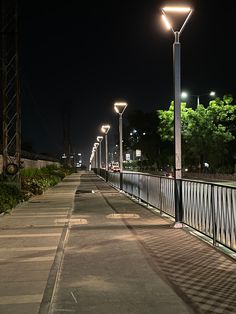 an empty sidewalk at night with street lights on the sides and trees in the background